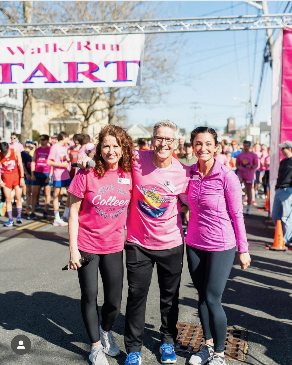 Three people smiling and posing at the start line of a walk/run event, with others behind them, wearing pink shirts.