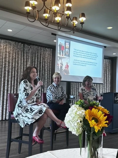 Three women seated on stage during a panel discussion at a conference or event, with a large screen behind them displaying text and images, and a flower arrangement with sunflowers in the foreground.