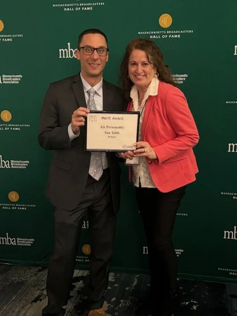 Two people standing in front of a green backdrop with 'Massachusetts Broadcasters Hall of Fame' and 'mba' logos. The man on the left is in a suit and glasses, holding a framed award. The woman on the right, wearing a red blazer, is also holding the a