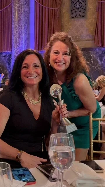 Two women smiling at a dinner event, one holding an award statue, in a decorated banquet hall with purple curtains and gold chairs.