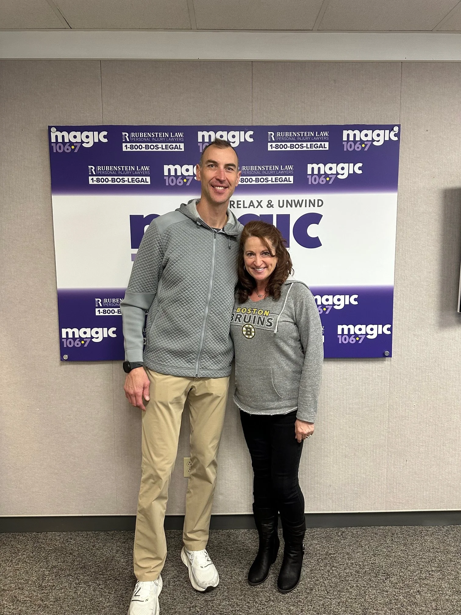 A man and a woman standing together indoors in front of a purple and white banner that promotes Magic 106.7 and Rubinstein Law. The man is wearing a gray jacket, khaki pants, and white sneakers, while the woman is dressed in a gray hoodie with Boston