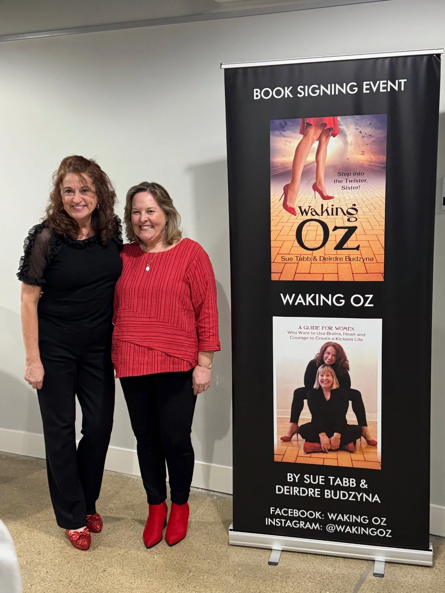 Two women standing next to a black book signing event banner for the book "Waking Oz" by Sue Tabb and Deirdre Budzyna. The woman on the left has curly brown hair, wearing a black top and black pants, and red high heels. The woman on the right has shoulder-length blonde hair, wearing a red top and black pants, and red high-heeled boots. Both are smiling.