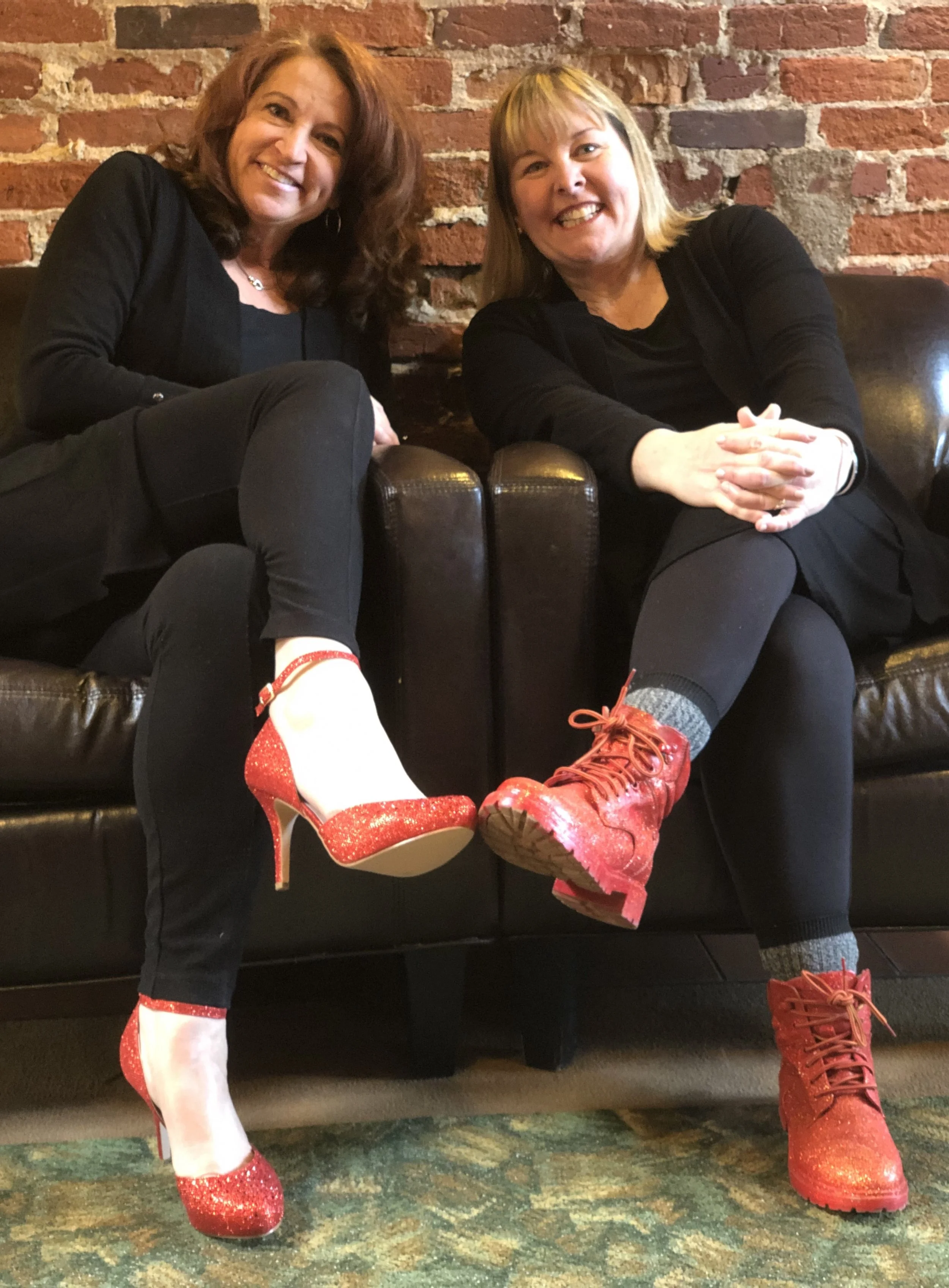 Two women sitting on a black leather couch against a brick wall, with one woman showing her red sparkly high heel shoe and the other showing her red sparkly boot, both smiling.