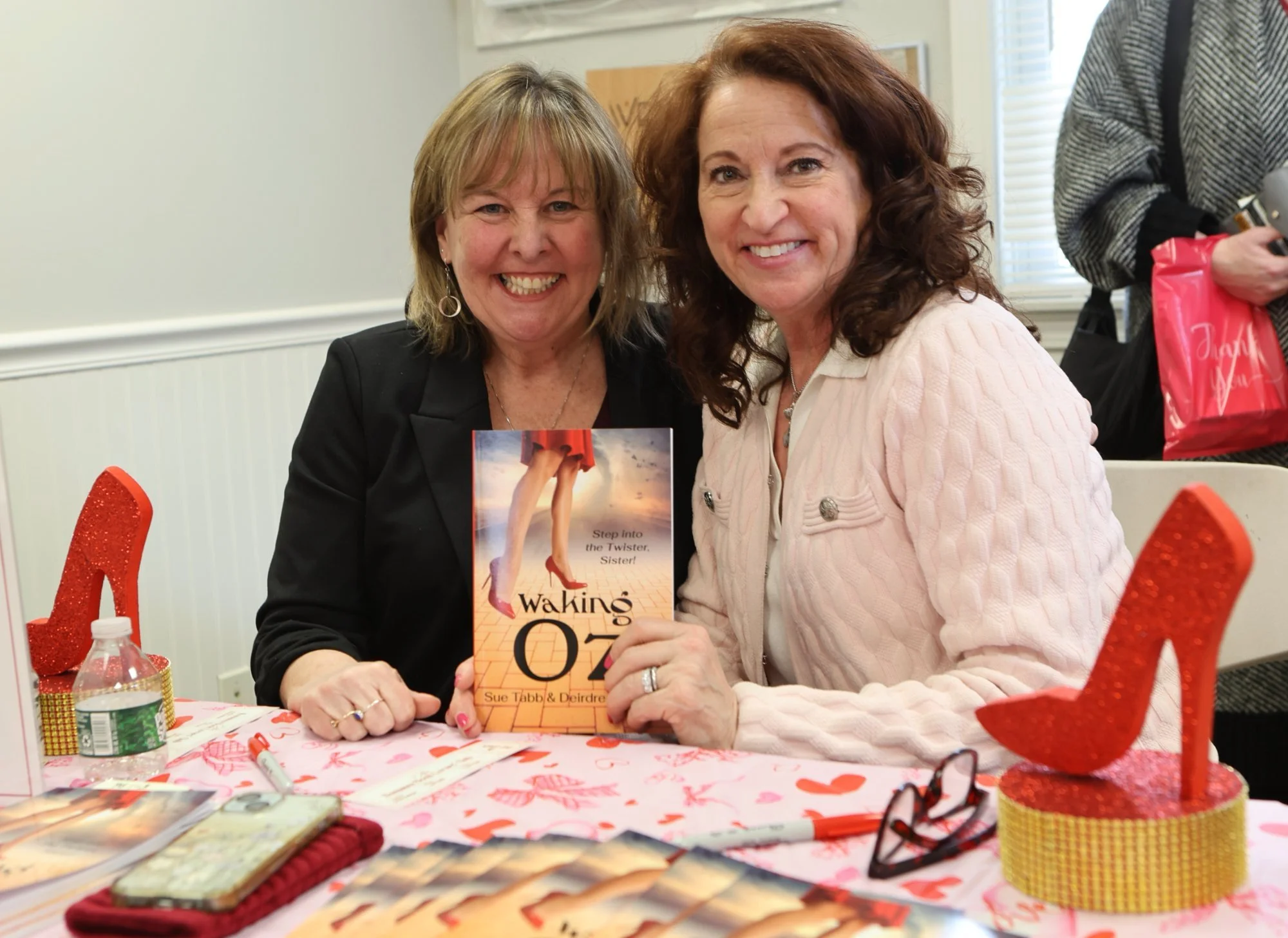 Two women smiling and holding a book titled "Waking O" at a Valentine's Day event, surrounded by Valentine's themed decorations, at a table with various items.