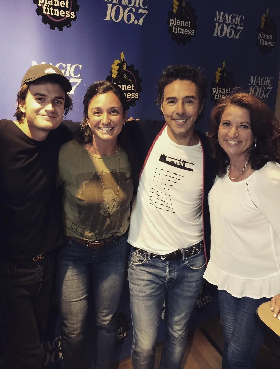 Group of four people smiling, standing close together in front of a blue backdrop with logos for Magic 106.7 and Planet Fitness.