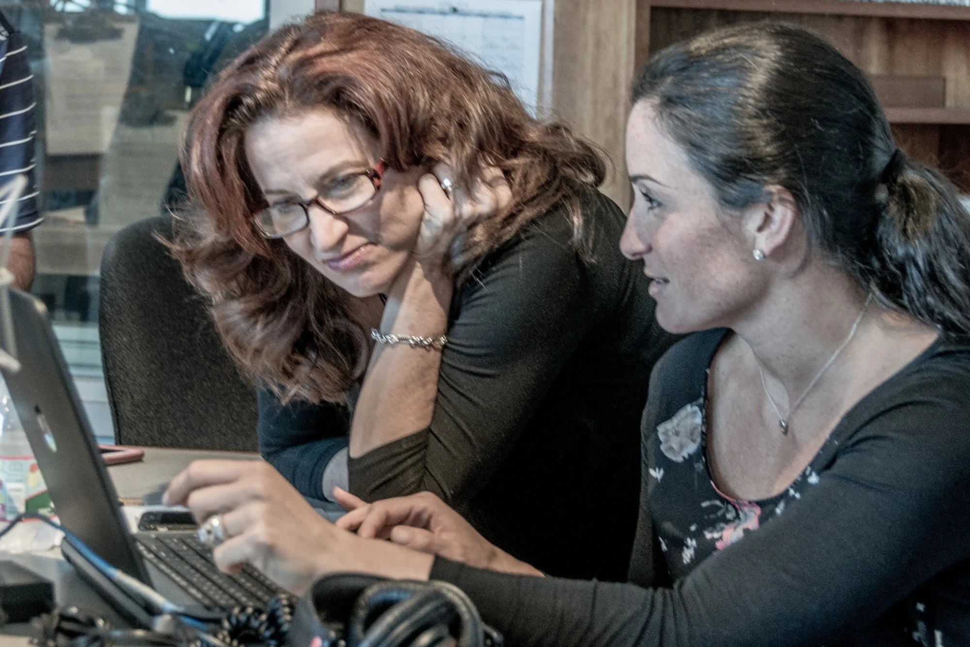 Two women are sitting at a table, looking at a laptop screen. The woman on the left has long, wavy red hair, glasses, and is resting her head on her hand, appearing thoughtful. The woman on the right has long dark hair tied back, is smiling, and appears to be explaining or discussing something with the other woman.