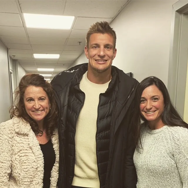 Three people standing together in a hallway, smiling at the camera. The person in the middle is a tall man wearing a black jacket over a cream sweater, with short hair. The woman on the left has curly brown hair and is wearing a textured cream-colore