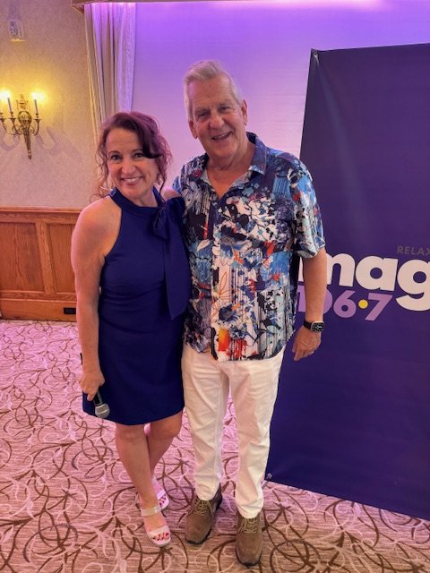 A woman in a blue dress and a man in a colorful Hawaiian shirt standing together indoors, smiling, with a purple banner and chandelier in the background.