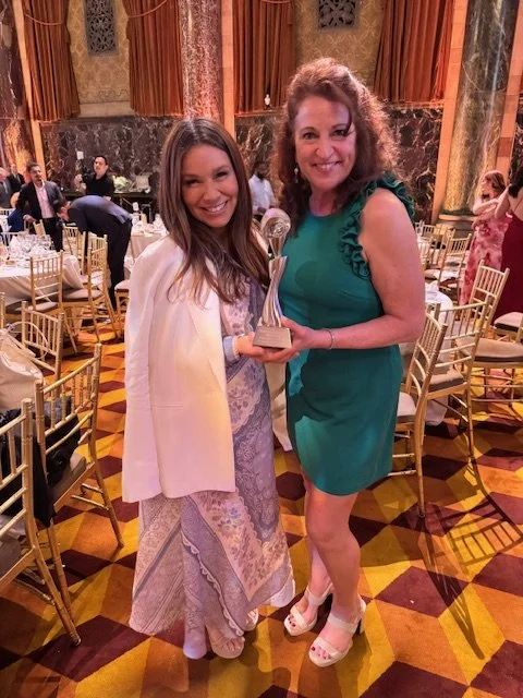 Two women are standing in a decorated banquet hall, holding a trophy and smiling. The woman on the left is wearing a long, patterned dress and a white cape. The woman on the right is wearing a green dress and white shoes. There are tables and chairs 