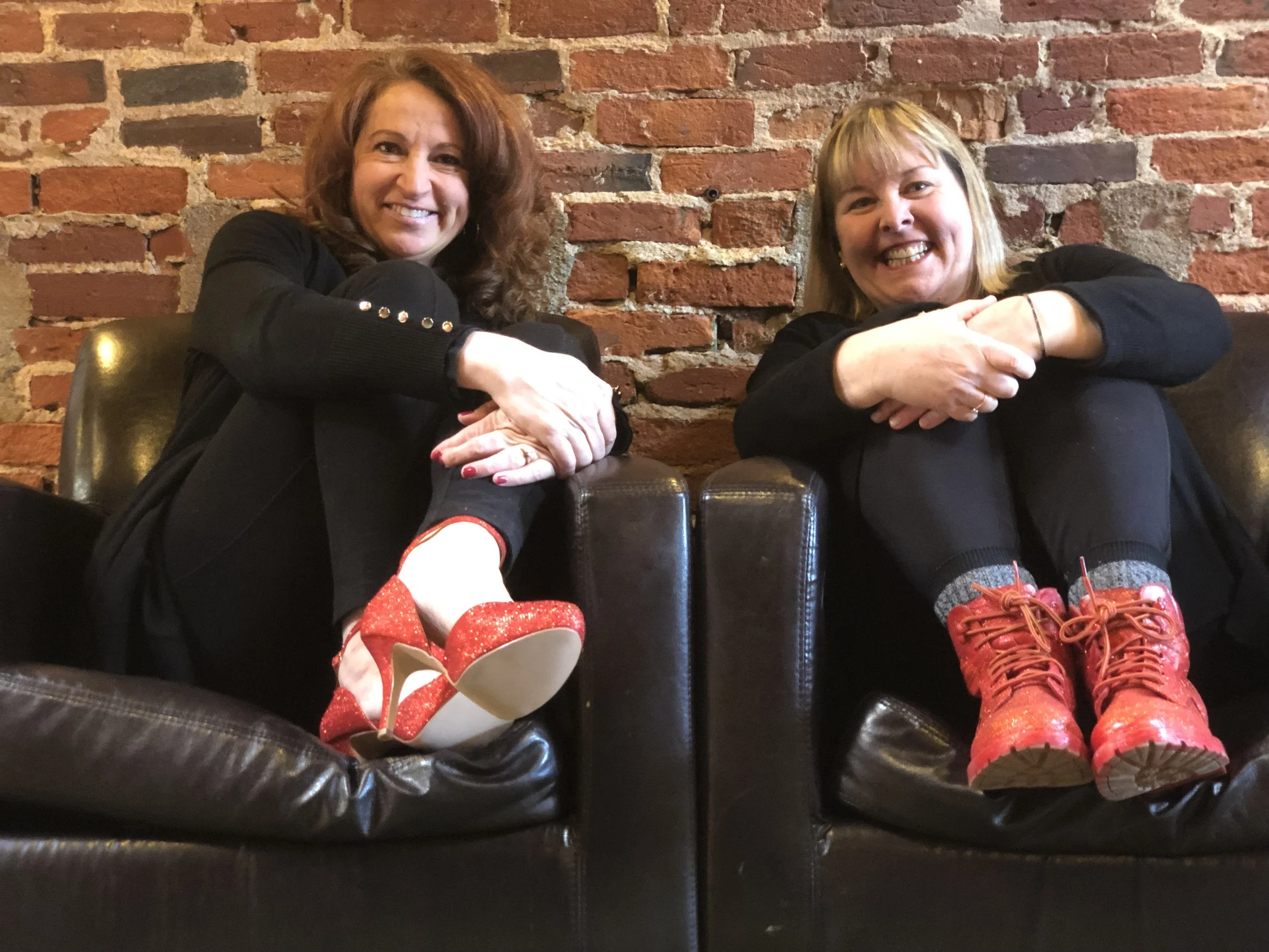 Two smiling women sitting against a brick wall, wearing black sweaters. The woman on the left has red hair, and the woman on the right has blonde hair. They are sitting with their arms resting on their knees, and are showing their legs with red shoes