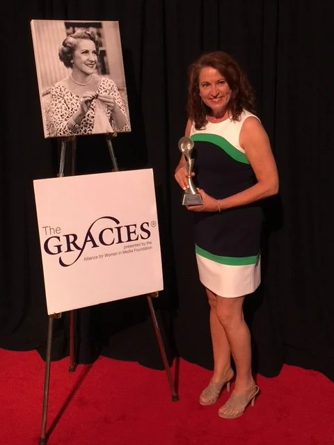 A woman standing on red carpet holding a trophy, next to a black and white photograph of herself, at The Gracies awards event.