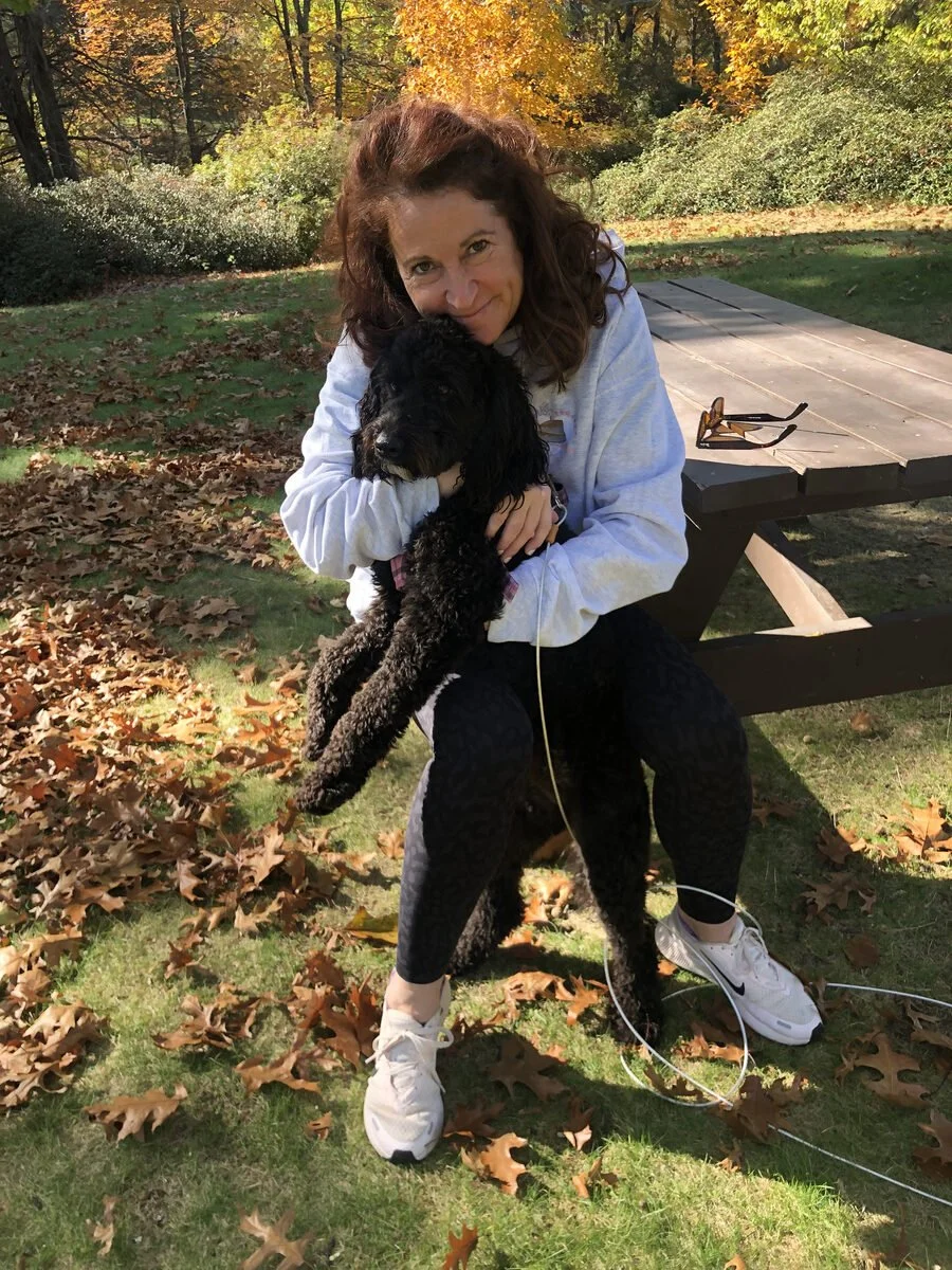 A woman with curly brown hair smiling while holding a black puppy outdoors in a park with autumn leaves on the ground and trees with orange and yellow foliage in the background. A picnic table with sunglasses on it is nearby.