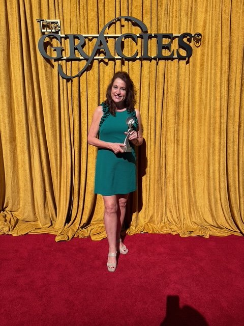 Woman in teal dress holding an award on stage at The Gracies award ceremony, with a gold curtain backdrop and The Gracies logo above.