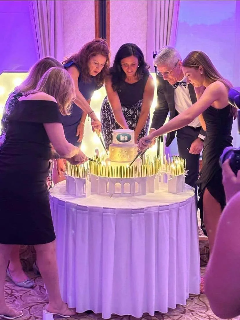 A group of six people, five women and one man, cutting a birthday cake together at a celebration. The cake has white frosting, with gold decorations and a small round topper. The scene is decorated with purple lighting.