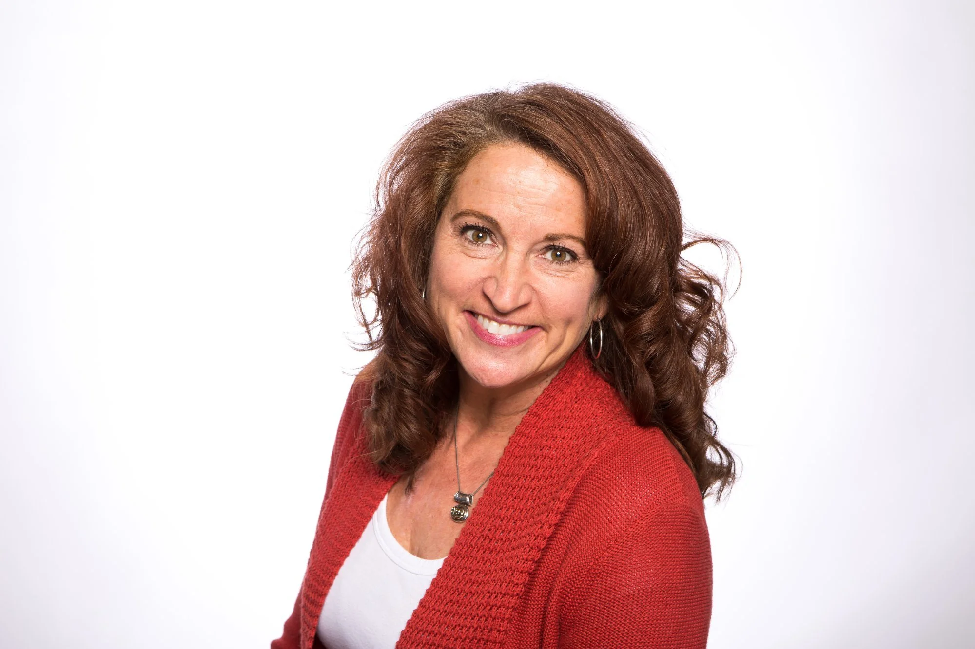 A woman with auburn, curly hair smiling at the camera wearing a red cardigan over a white top, with a silver necklace and hoop earrings, against a plain white background.