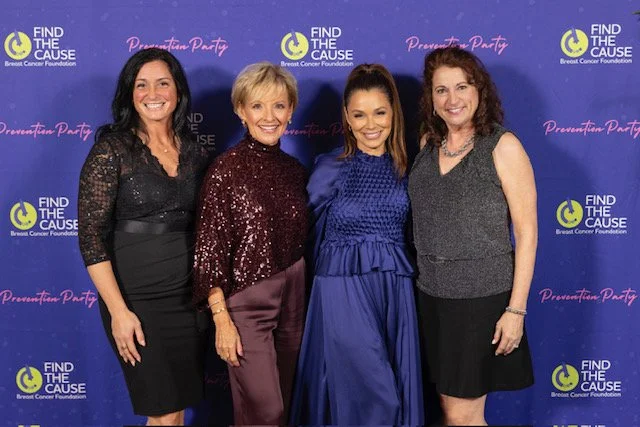 Four women standing together in front of a blue backdrop with the logos of the Breast Cancer Foundation and the Find the Cause campaign, smiling for the camera at a formal event.