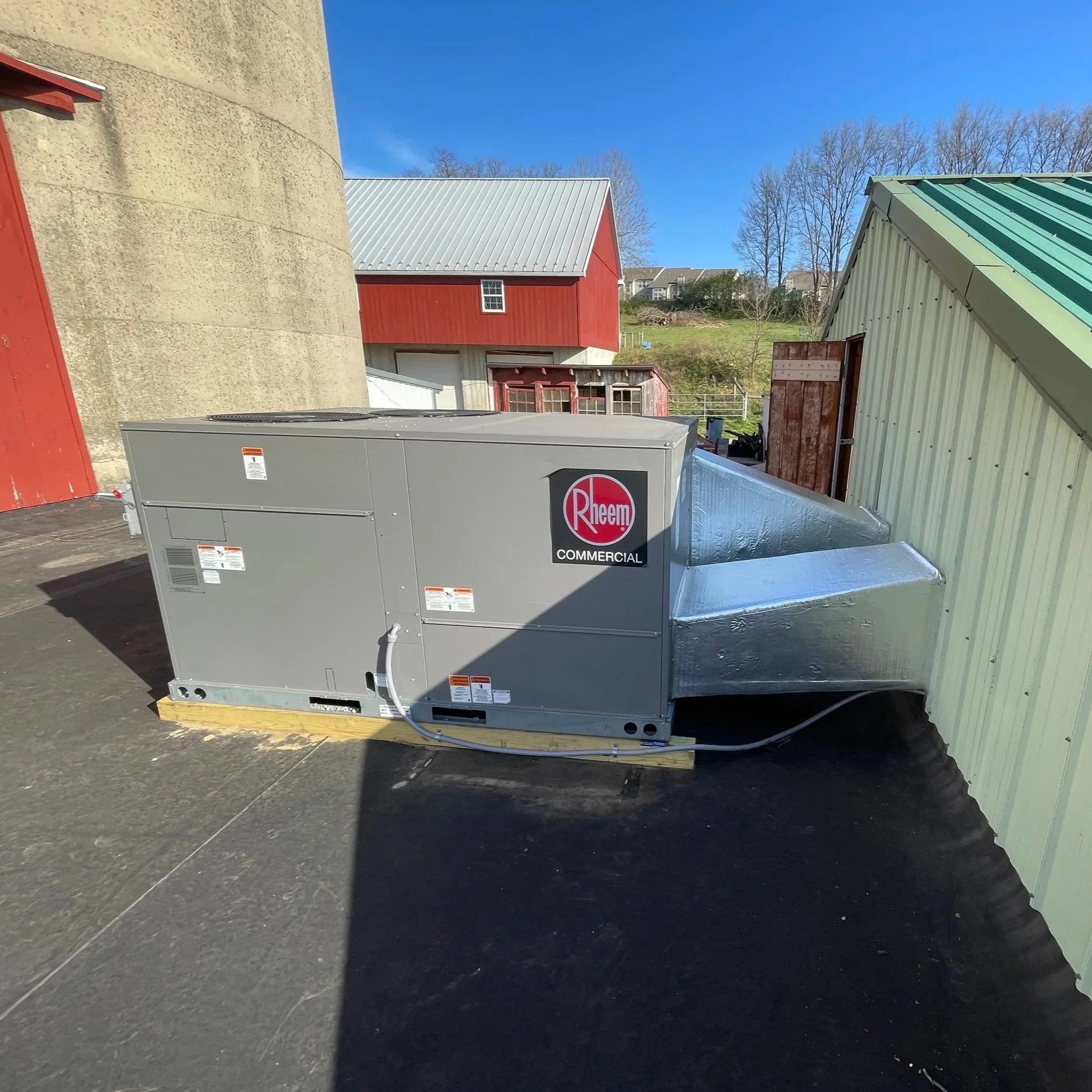 An industrial HVAC unit with a Rheem label, installed on a wooden pallet outside near buildings with metal roofs, against a background of leafless trees and a blue sky.