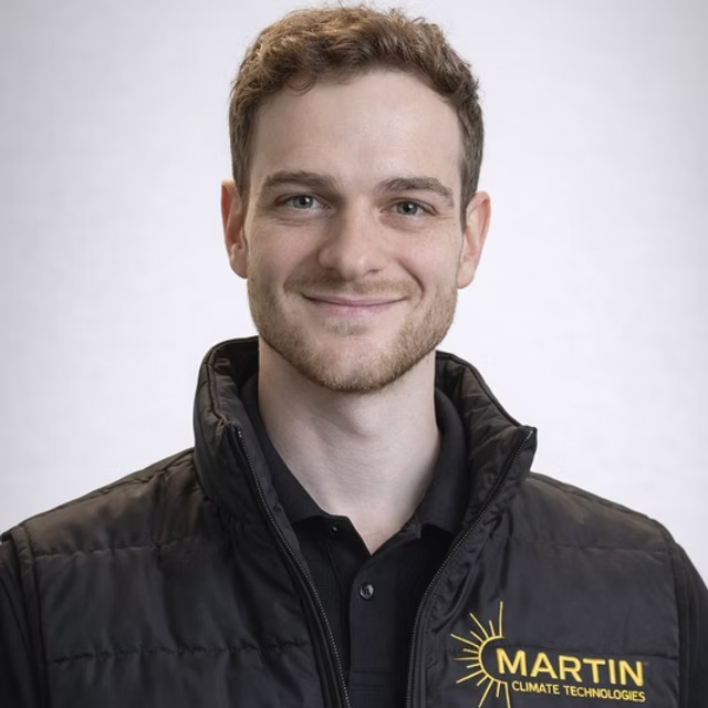 A young man with light brown hair and a short beard smiling at the camera, wearing a black jacket with a yellow logo that says "MARTIN CLIMATE TECHNOLOGIES" on the chest, against a plain gray background.