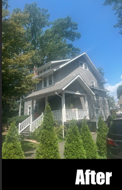 A gray, multi-story house with a front porch, surrounded by well-maintained shrubs, under a clear blue sky with trees nearby.