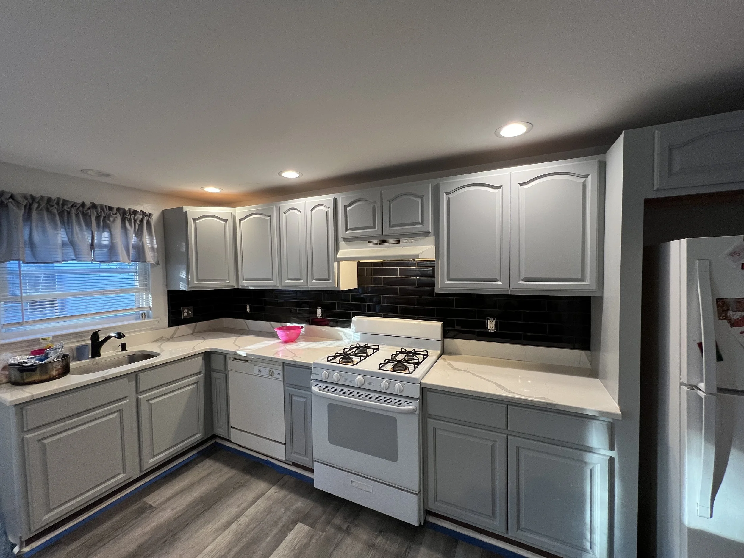 Kitchen with white cabinets, a window with a ruffled curtain, and a white stove and oven. Countertop with pink bowl, faucet, and various kitchen items. Dark backsplash, hardwood floor, and overhead lighting.