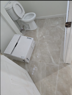 A small bathroom with a white toilet, a cardboard box on the floor, and beige tiled flooring.