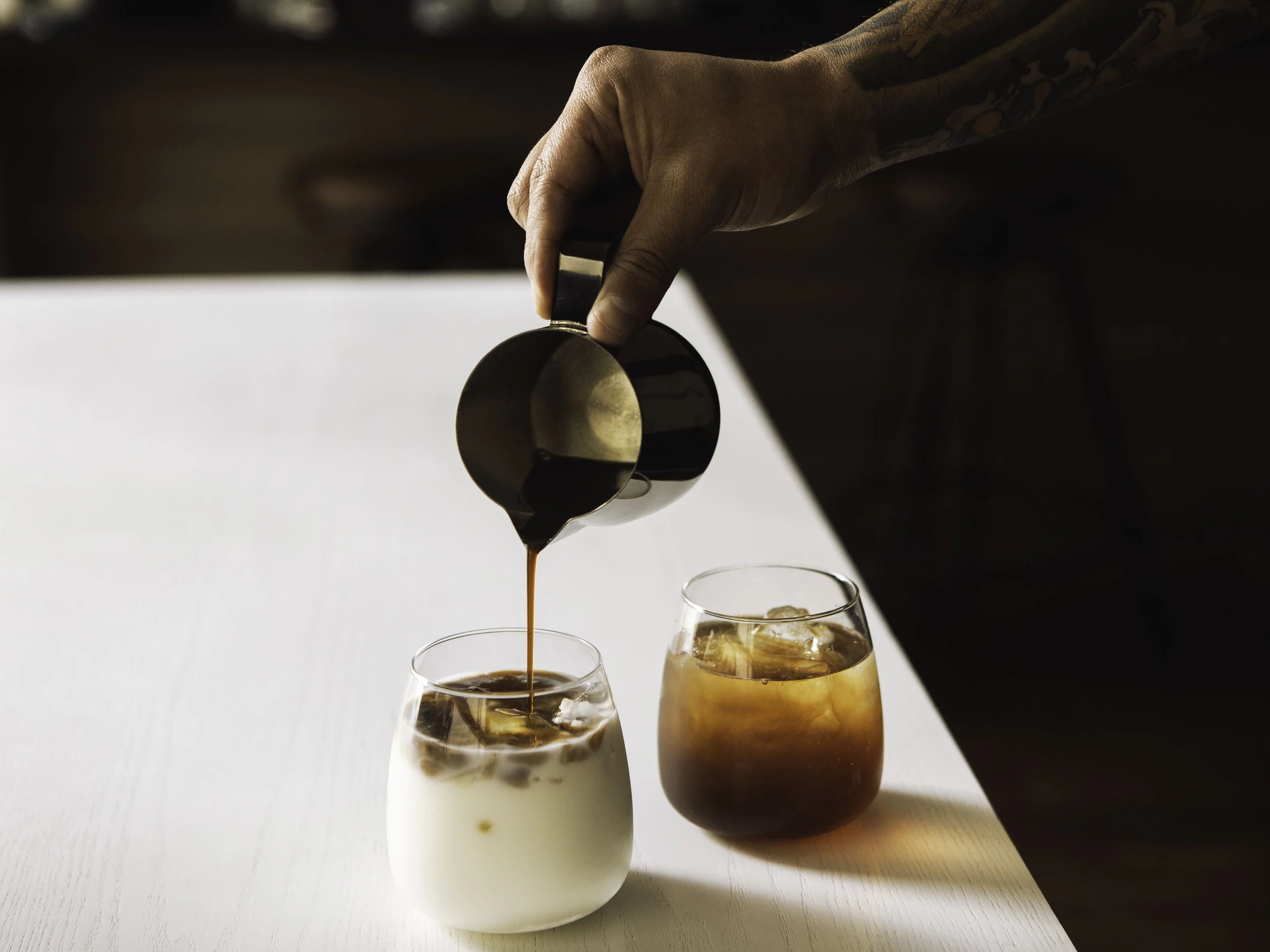 A hand pours dark coffee from a small pitcher into a glass with milk and ice. Another glass with iced coffee is next to it on a white surface.