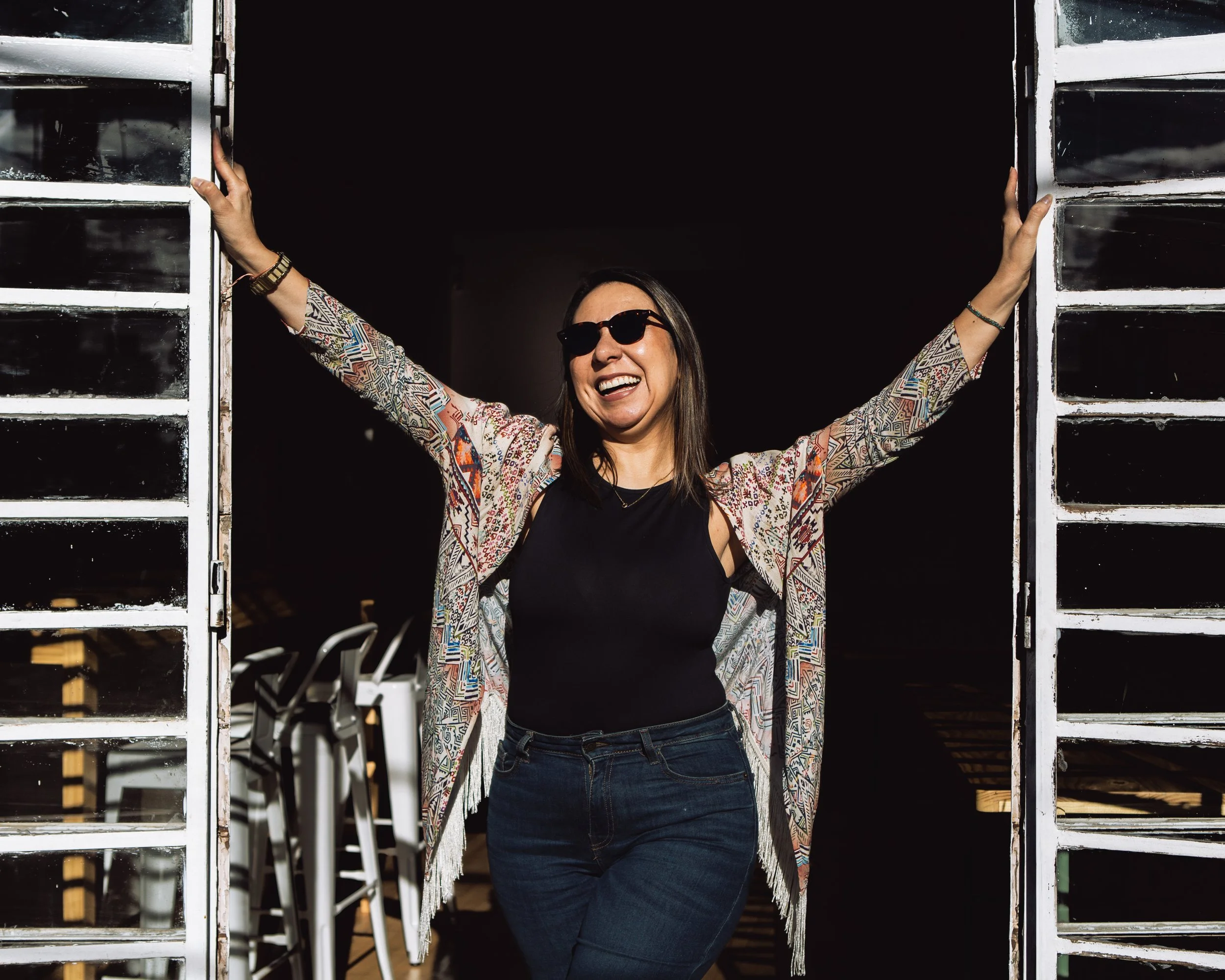 Woman smiling and celebrating with arms raised, standing between open doors, wearing sunglasses, a black top, and a colorful patterned jacket.