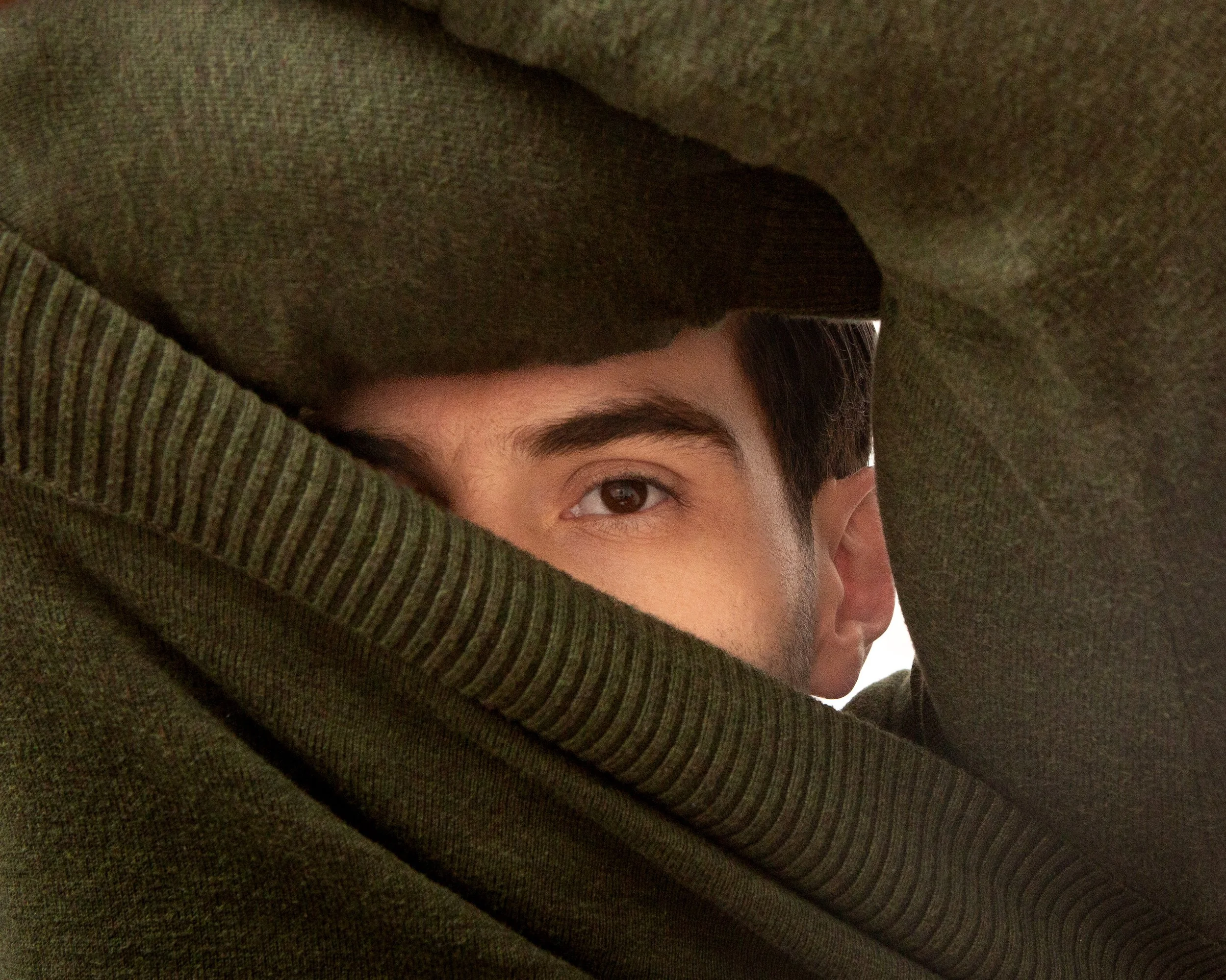 A person peeking through a small opening in a green fabric, revealing one eye and part of their face.