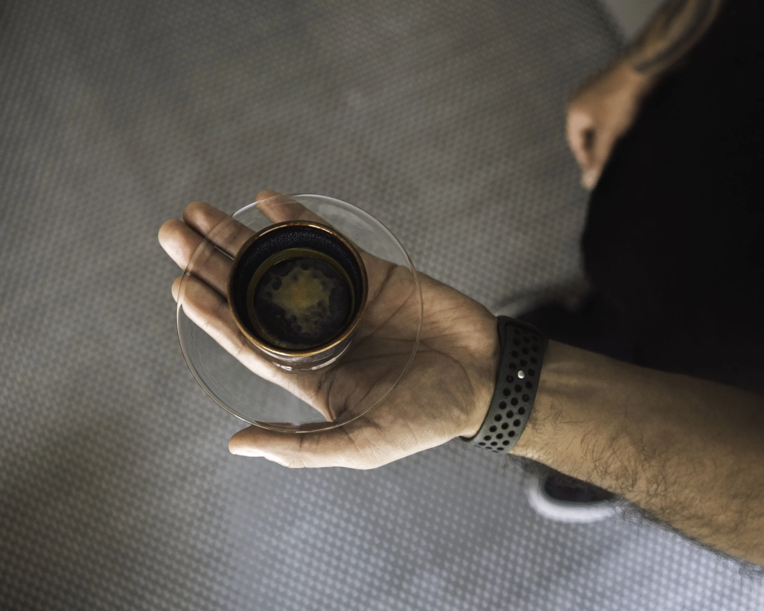Person holding an espresso in a glass cup, viewed from above, with a patterned gray surface in the background.