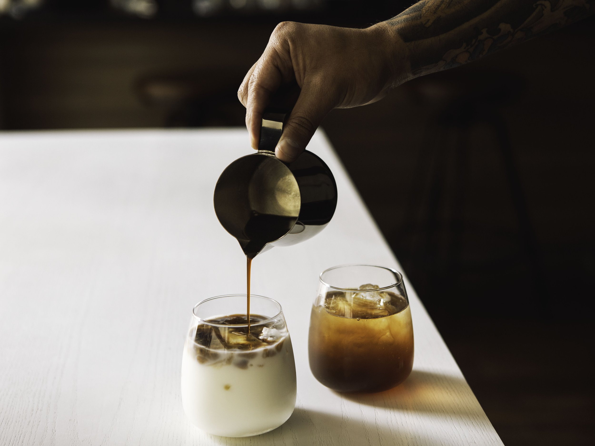 Person pouring coffee from a stainless steel pitcher into a glass of iced coffee with milk, with another glass of iced coffee on the side.