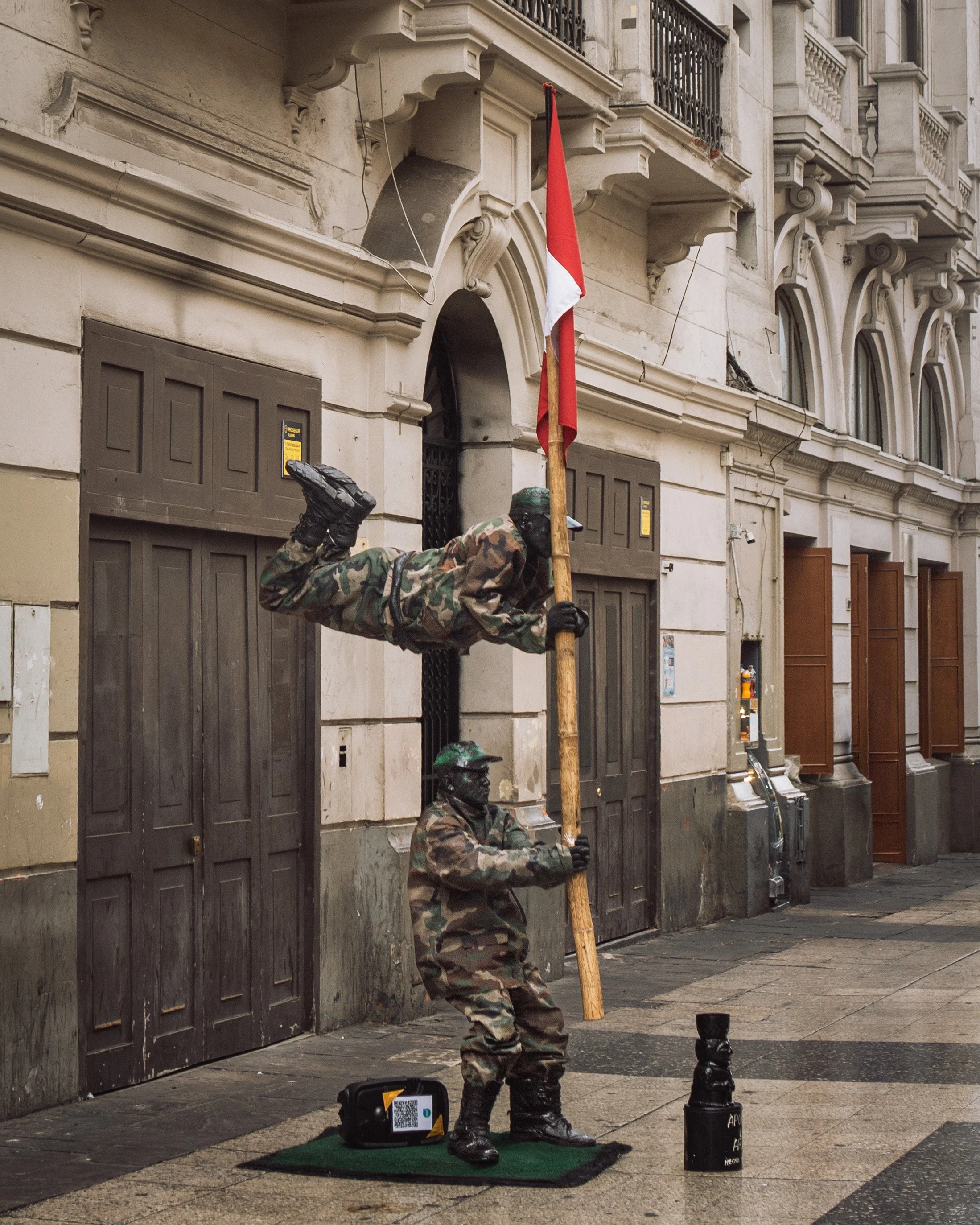 Two statues of soldiers in camouflage uniforms performing a stunt with one holding a flag, mounted on a street sidewalk in front of a building.