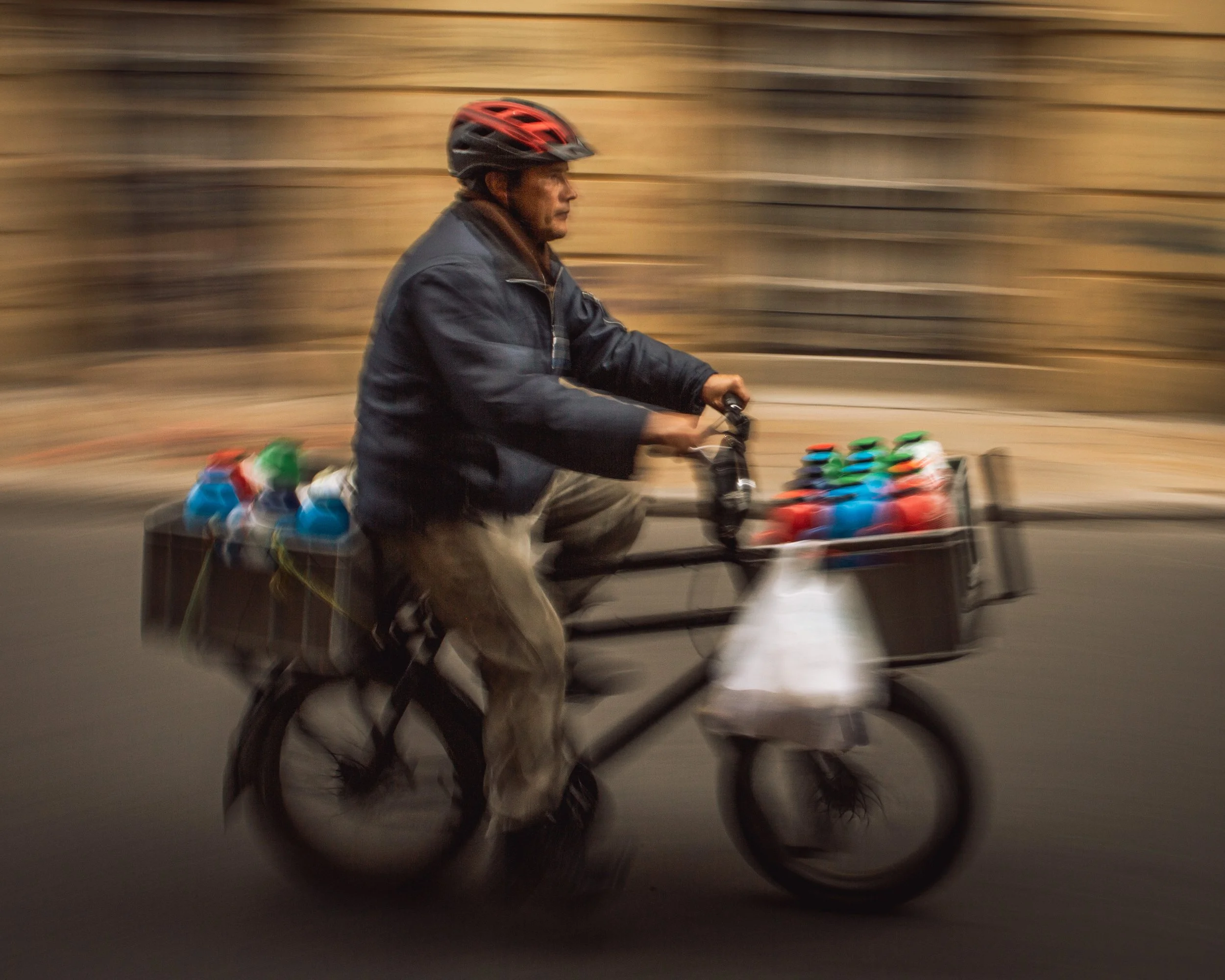 A man riding a bicycle with two baskets filled with colorful bottles, blurred background indicating motion, wearing a black helmet and dark jacket.