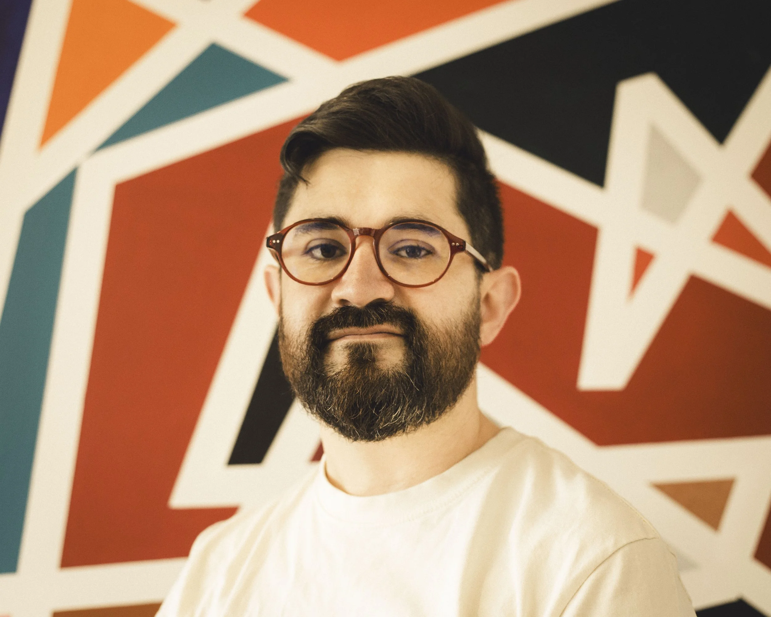 Portrait of a man with glasses and a beard, standing in front of a colorful geometric mural.