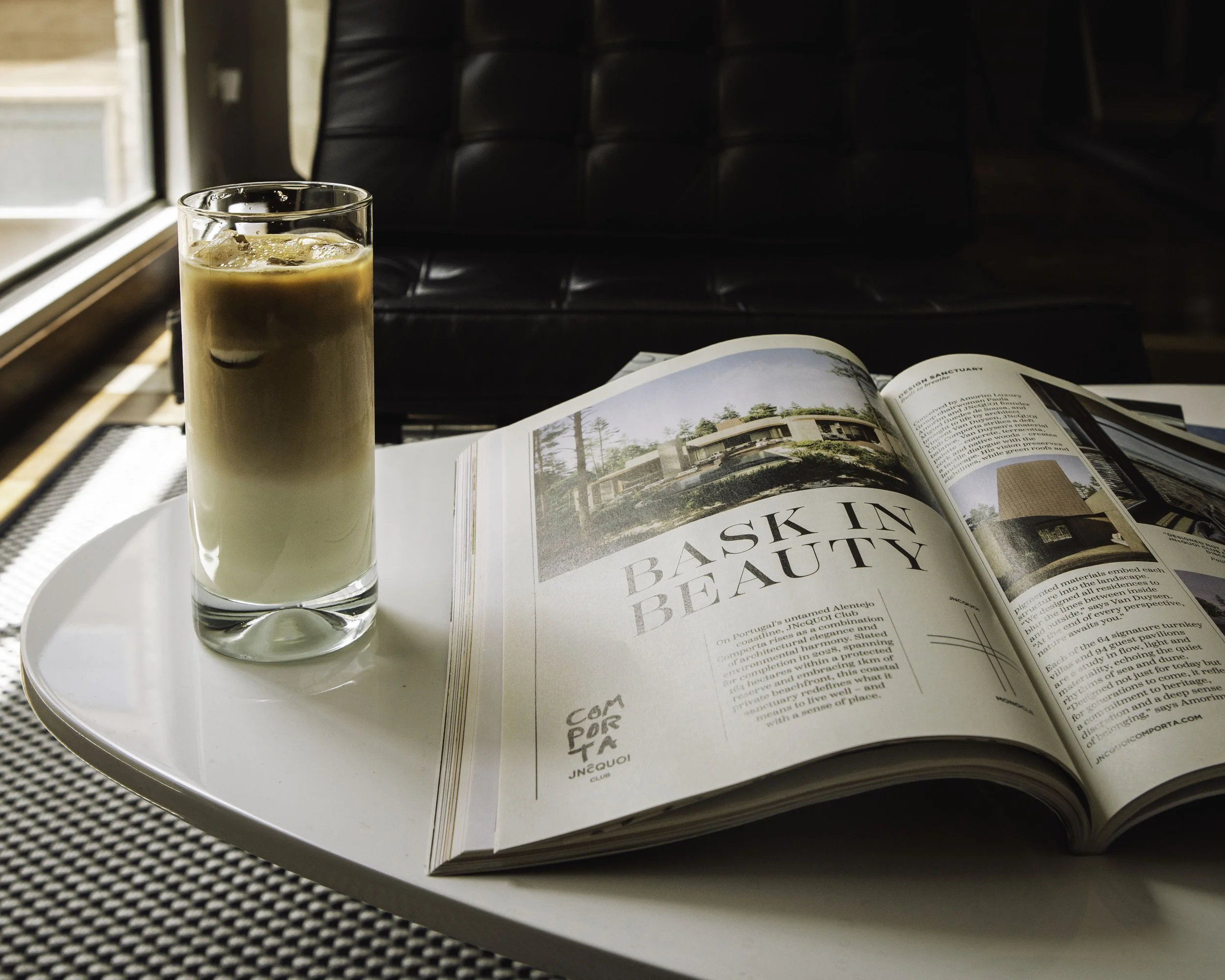 A glass of iced coffee and an open magazine on a white table near a window with a black chair in the background.