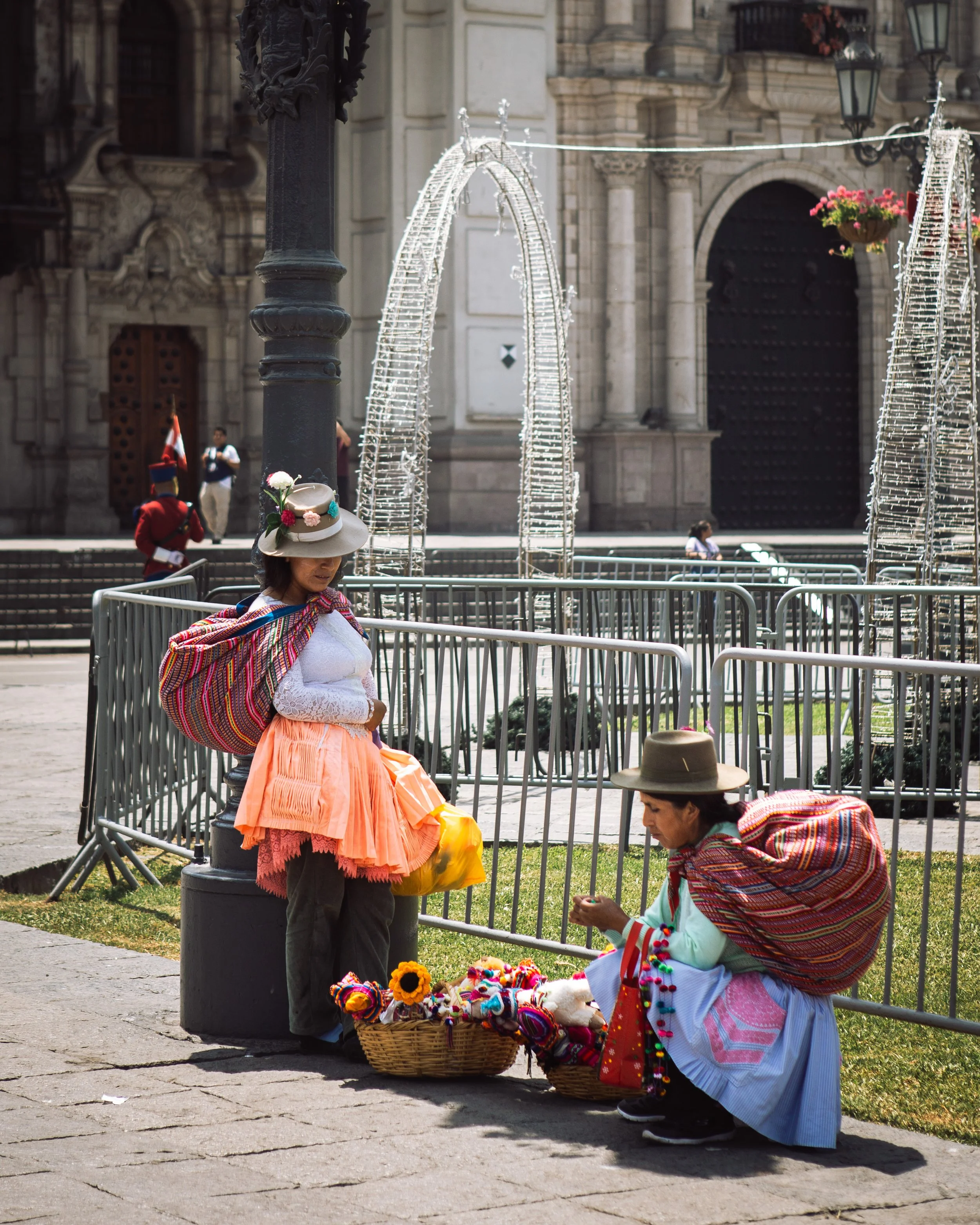 Two women in traditional colorful clothing and hats selling handmade crafts, such as flowers and jewelry, on a city street with a historic building in the background.