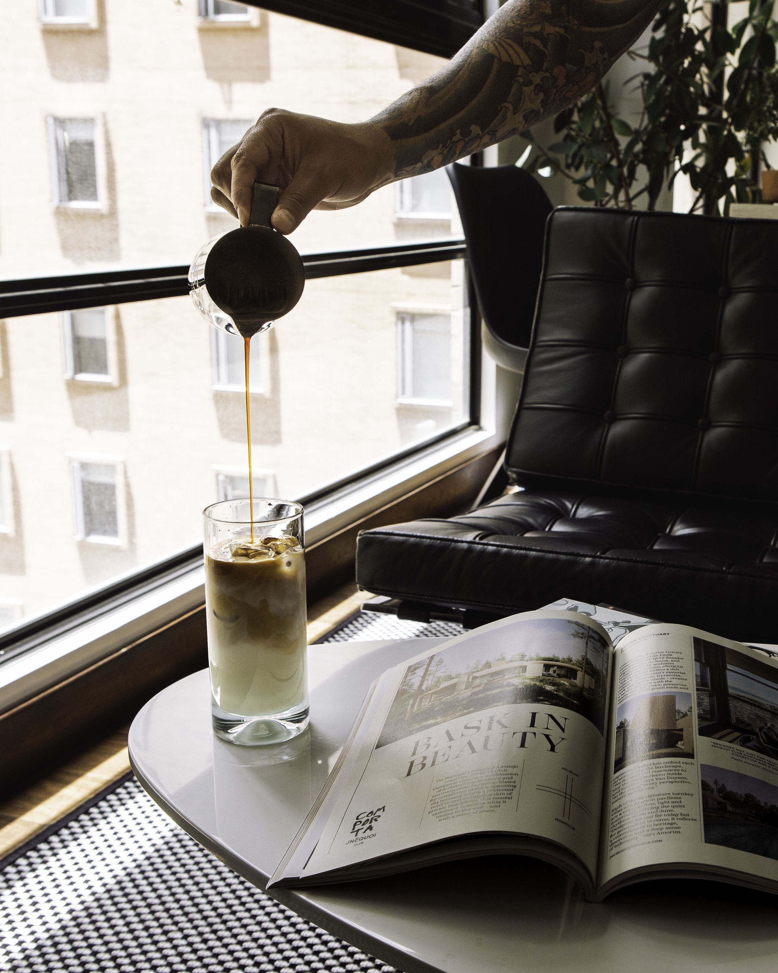 A person with tattoos on their arm pouring coffee into a glass of iced coffee on a white table, with an open magazine nearby, beside a black leather chair near a window.