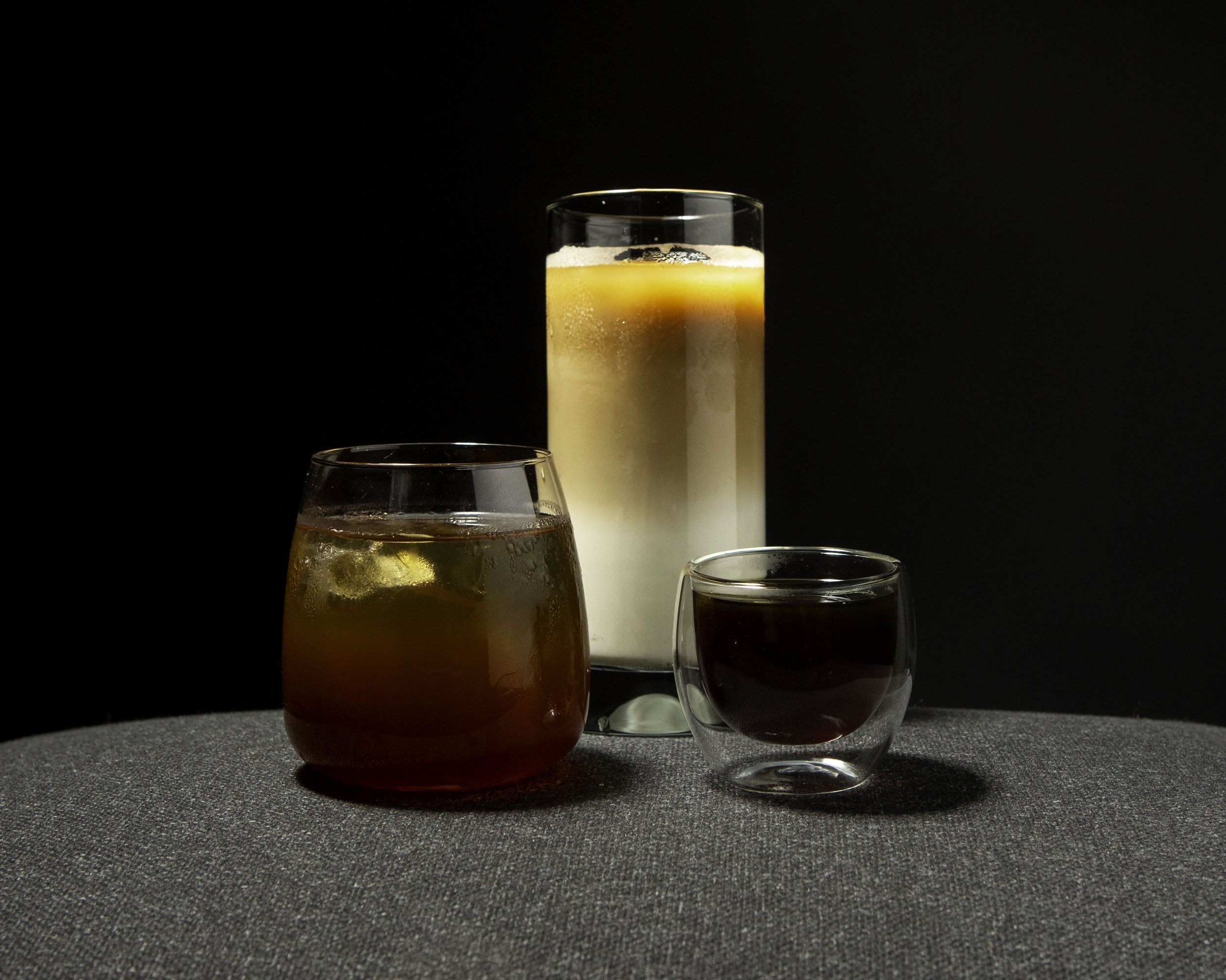 Three different glasses with beverages on a textured gray surface against a black background. The left glass contains a dark-colored drink with ice, the middle glass has a layered drink with a yellow top and white bottom, and the right glass holds a 