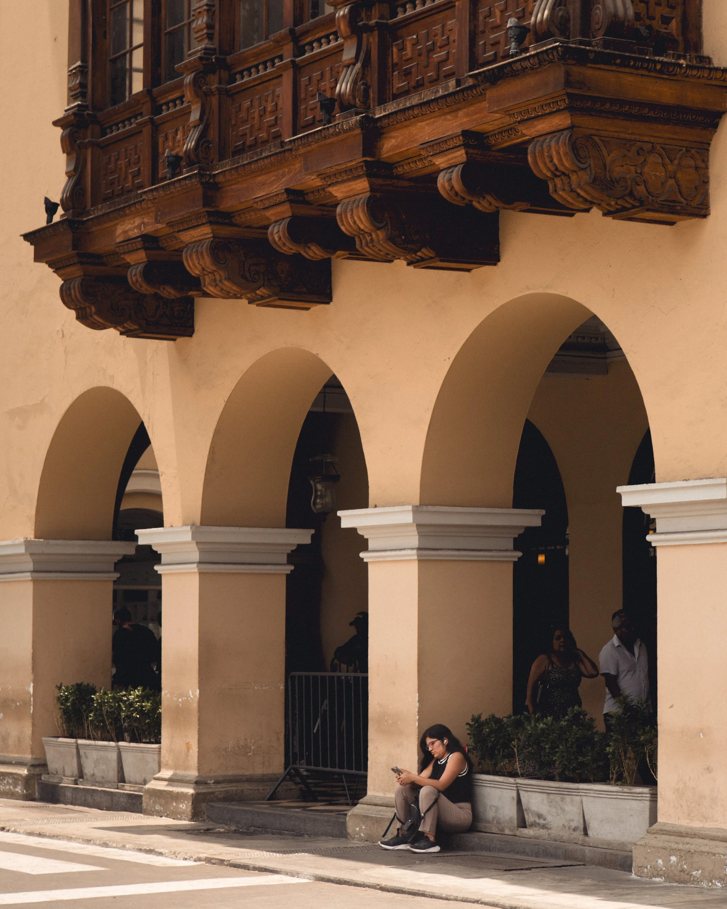A woman sitting on the sidewalk and looking at her phone outside a colonial-style building with arched openings and a decorative wooden balcony.