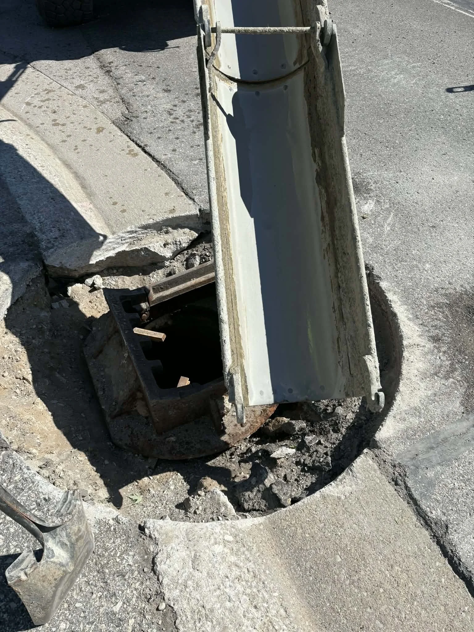 Open manhole with sidewalk around it, construction work in progress, concrete being poured by heavy equipment.