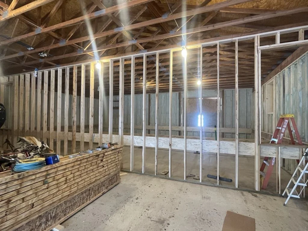 Interior view of a building under construction with a wooden framed wall, exposed ceiling joists, and construction tools and materials nearby.