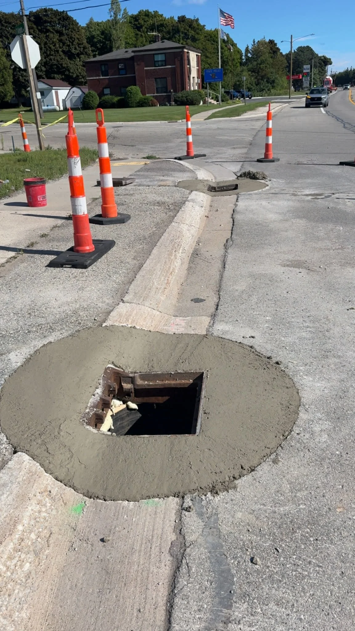 A construction site on a city street shows a square hole in the ground with fresh concrete around it and orange traffic cones surrounding the area.