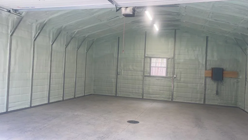 Empty garage with metal framing, spray foam insulation, concrete floor, small window, utility box on wall.