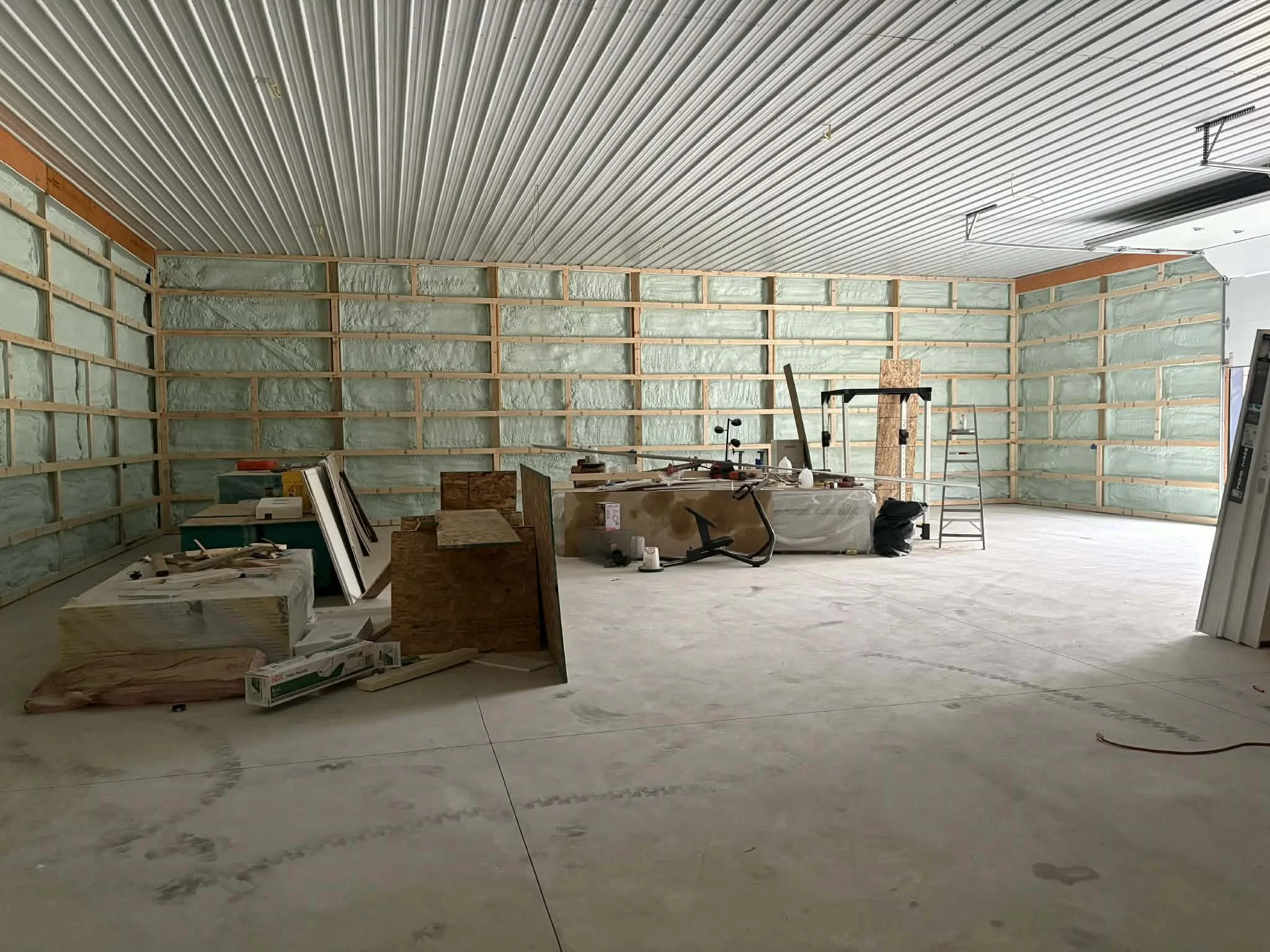 Interior of a room under construction with spray foam insulation on the walls, building materials, tools, and a ladder scattered around.