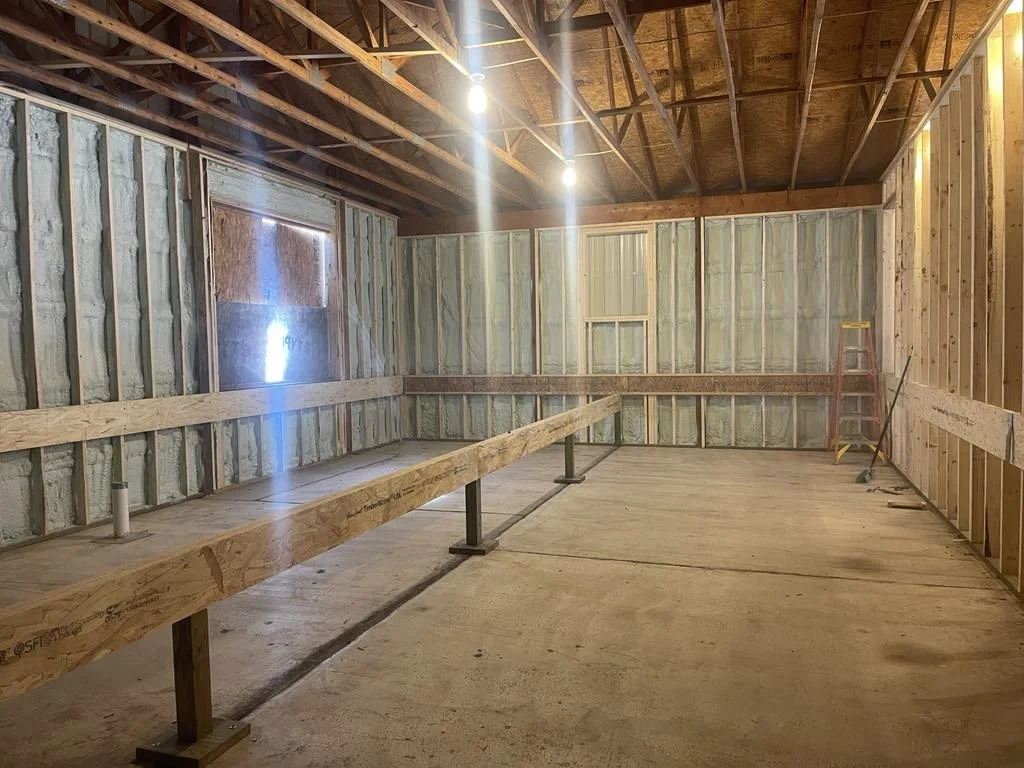 Interior of a garage under construction with exposed wooden framing with spray foam insulation, a concrete floor, a ladder in the corner, and two bright ceiling lights.