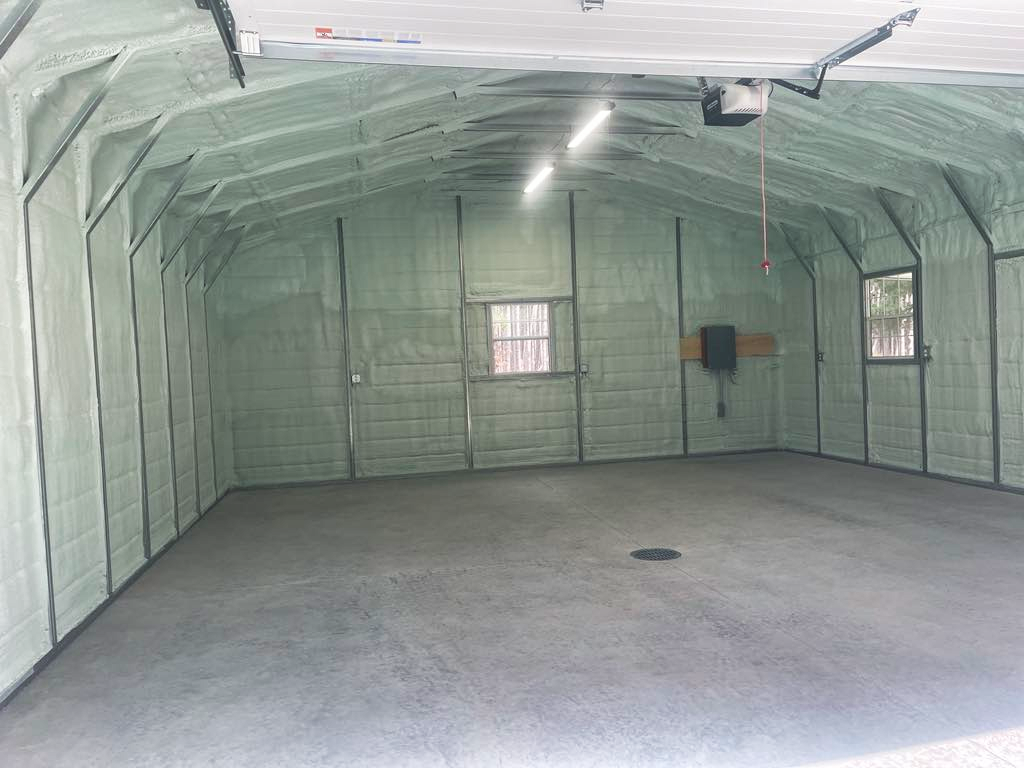 Empty garage or storage shed with spray foam insulated walls, two windows, overhead lights, and a garage door.