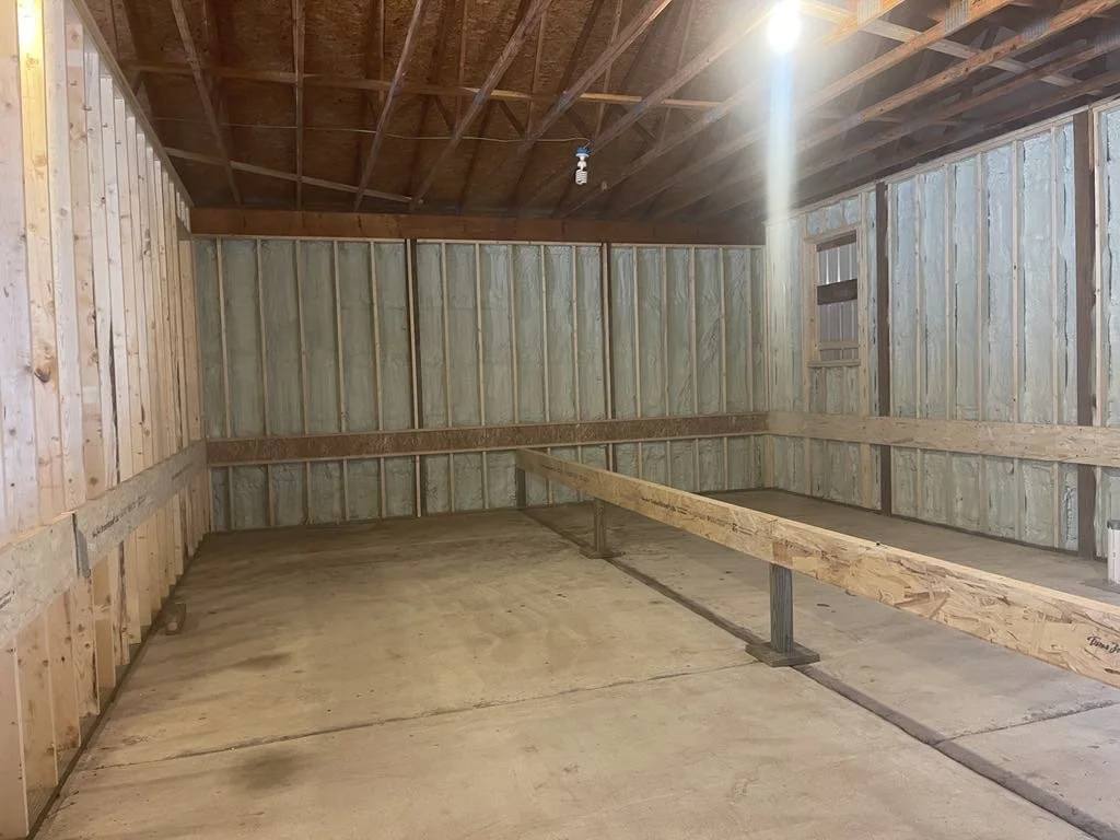 Empty unfinished garage with wooden framing, spray foam insulation, a concrete floor, and a single light bulb hanging from the ceiling.