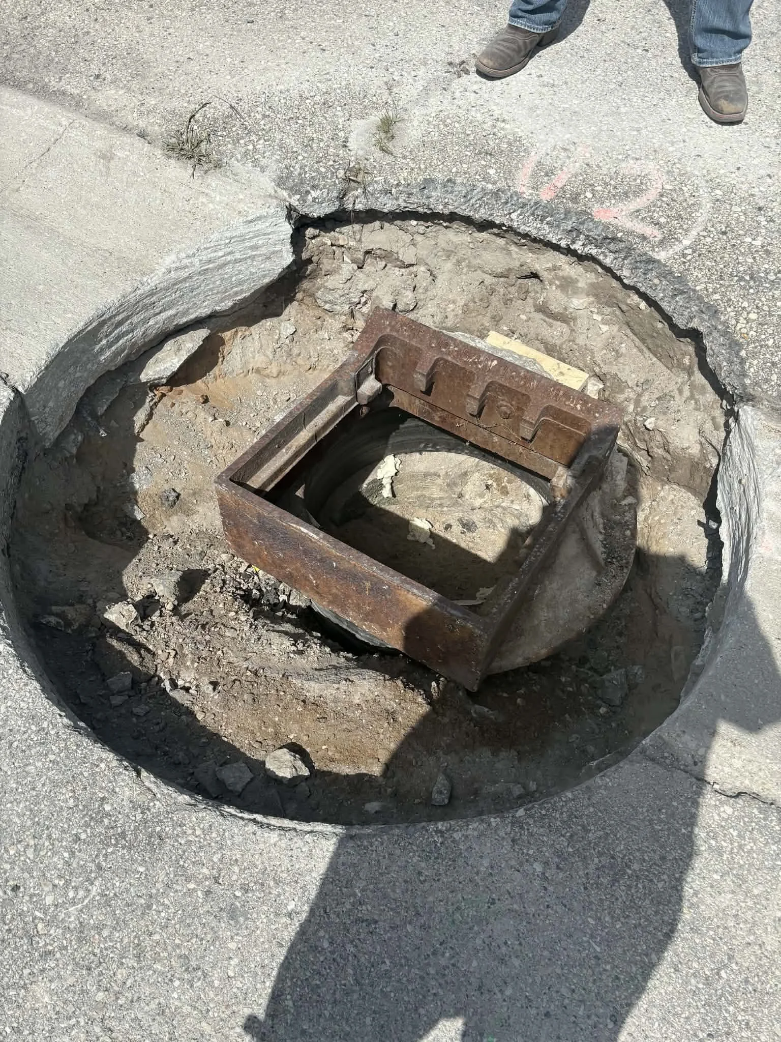 An exposed storm drain, exposing the inner plumbing, with an open circular asphalt cover and a person standing nearby.