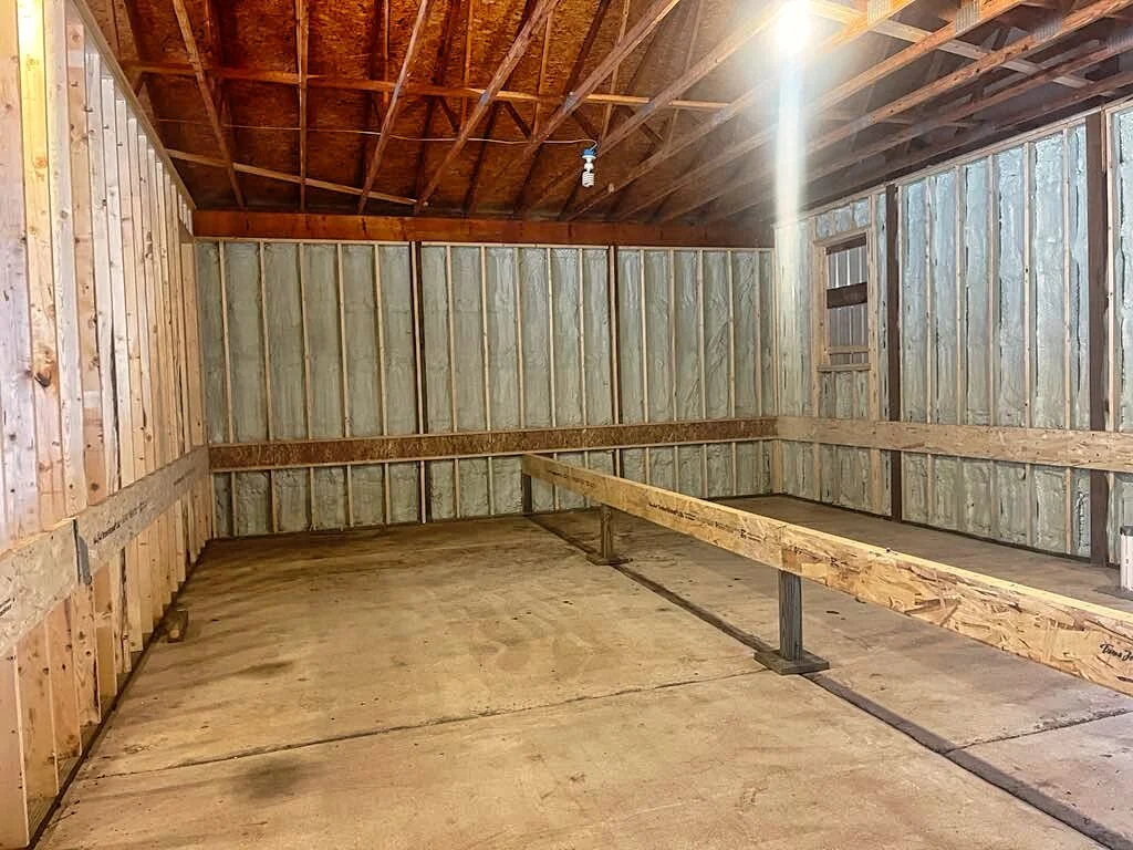 An unfinished garage with wooden framing, spray foam insulation, exposed roof trusses, and a concrete floor. There is a vertical window on the right wall and a small light fixture hanging from the ceiling.