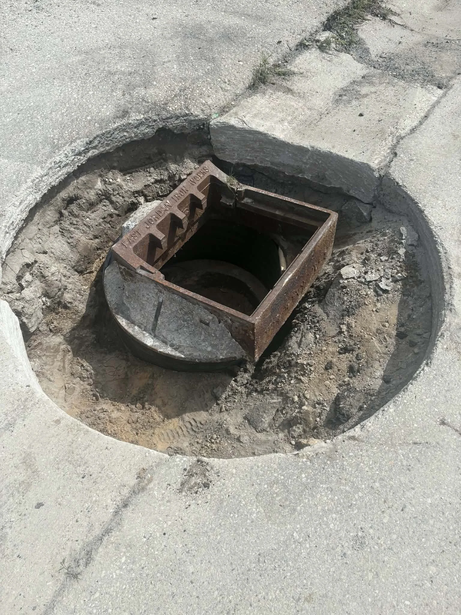 An open metal access cover or manhole in a concrete sidewalk, exposing a dark interior and marked with the words 'West Junction Iron Works'.