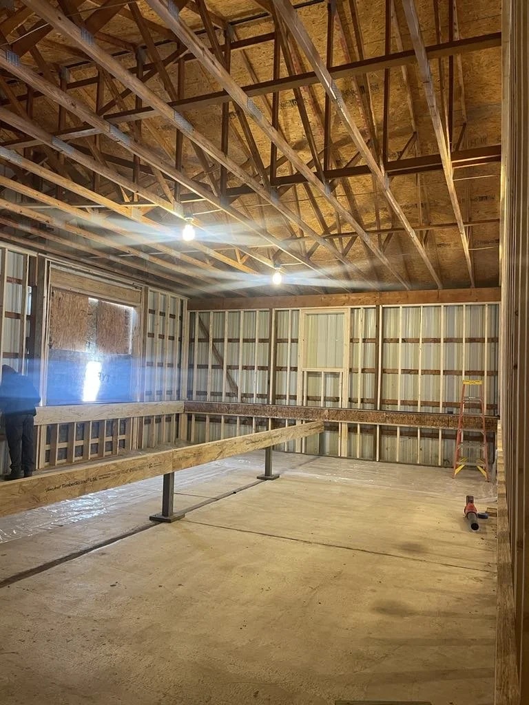 Interior of a building under construction, showing exposed wooden framing, spray foam insulation, a plywood ceiling, and a partially built railing around the perimeter.